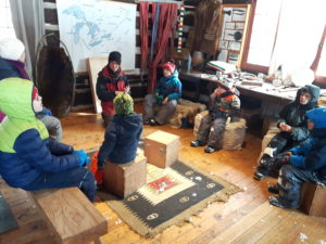 Photo of kids in a winter cabin, learning about traditional nature skills from a teacher.